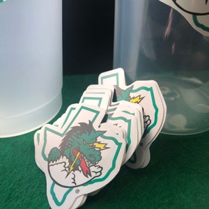 May include: A stack of green, white, and yellow stickers featuring a cartoon dragon with a lightning bolt in its mouth. The dragon is inside the outline of the state of Texas. The stickers have a registered trademark symbol.