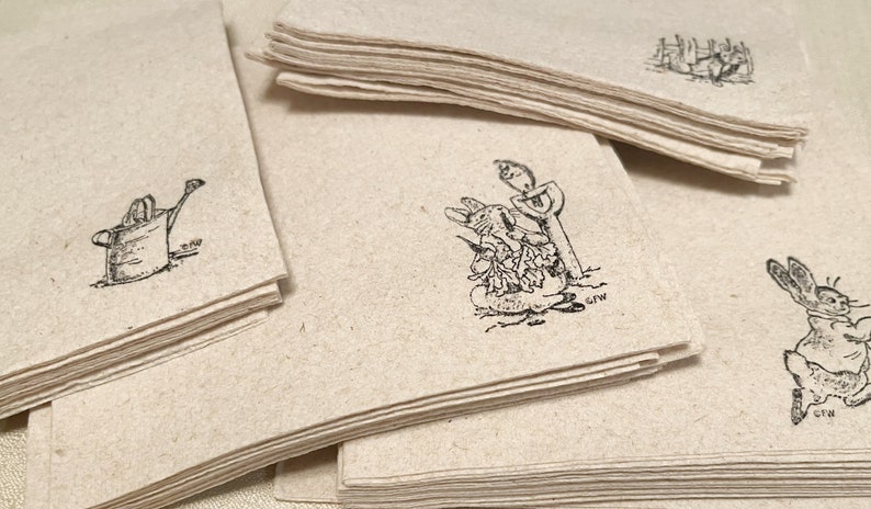 Pode incluir: Uma pilha de cart&otilde;es de papel bege com ilustra&ccedil;&otilde;es em tinta preta de coelhos e ferramentas de jardinagem. As ilustra&ccedil;&otilde;es incluem um regador, um coelho com uma p&aacute; e um coelho perto de uma cerca. Os cart&otilde;es t&ecirc;m uma apar&ecirc;ncia r&uacute;stica.