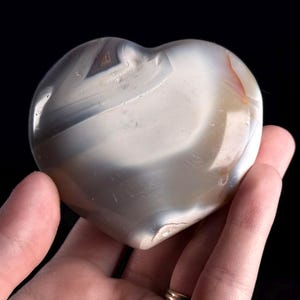 May include: A heart-shaped stone with a polished surface. The stone features a mix of white, gray, and brown bands and swirls. The heart is held in a hand against a black background.