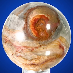 Può includere: Una gemma sferica lucidata con un motivo a spirale di colori. L'orbita presenta un'area centrale di rosso scuro e marrone, circondata da tonalità più chiare di beige, crema e grigio. La pietra è traslucida, permettendo alla luce di passare.