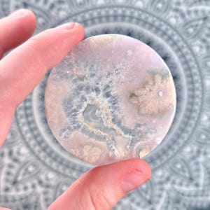 May include: A circular polished stone slice featuring a pale pink and translucent white color palette. The surface displays intricate crystalline formations, natural mineral inclusions, and a central jagged cavity with textured edges.