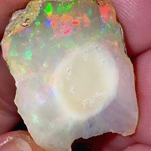 May include: A rough-cut opal gemstone with a creamy white base and vibrant flashes of color, including red, green, and blue. The opal has a natural, irregular shape and a polished, central area. The gemstone is a unique piece of natural jewelry.