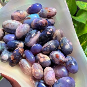 May include: A collection of polished, oval-shaped purple agate stones with varied inclusions. The stones feature shades of lavender, deep violet, and soft pink, with some displaying intricate, mottled patterns and translucent, smooth surfaces.