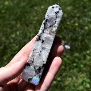 May include: A polished, hexagonal crystal tower featuring a translucent white and grey base with prominent black mineral inclusions throughout the stone. A small, iridescent blue flash is visible near the bottom edge of the faceted mineral point.