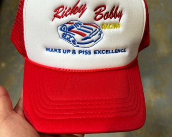 May include: Red and white trucker hat with the text "Ricky Bobby Racing" and a stylized race car graphic. The hat also features the phrase "Wake up & piss excellence" in blue. The hat has a red brim and mesh back.