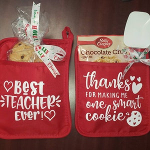 May include: Two red oven mitts with white text. The left mitt says "Best Teacher Ever!" and has a chocolate chip cookie in the pocket. The right mitt says "Thanks for making me one smart cookie" and has a bag of chocolate chip cookie mix and a spatula.