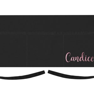 May include: Black apron with three pockets and adjustable straps. The apron has the name "Candice" embroidered on the front in pink.