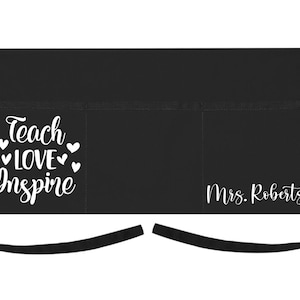 May include: Black apron with two pockets and adjustable straps. The apron has white text that says "Teach Love Inspire" with hearts around the words. The text "Mrs. Roberts" is also printed in white on the apron.