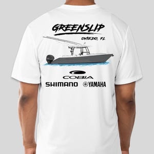 May include: White t-shirt with a black graphic of a boat, the text "GREENSLIP OVIEDO, FL", and the logos "COBIA", "SHIMANO", and "YAMAHA".