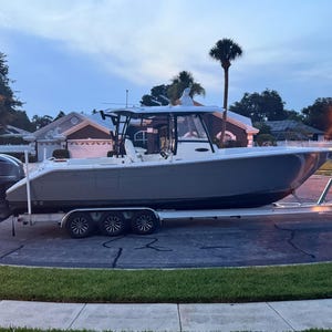 May include: A gray and white boat on a trailer with a white, black, and gray engine. The boat has a hardtop with a windshield and a gray hull. The boat is parked on a paved driveway in front of a house.