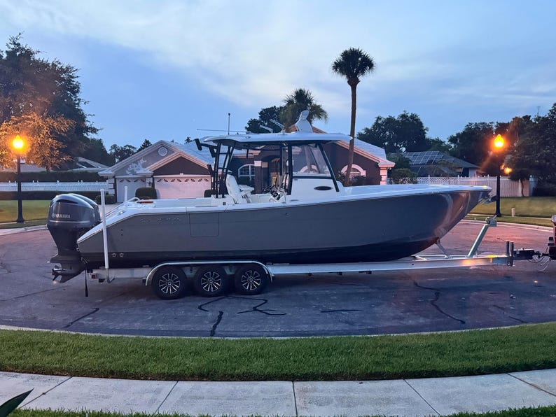 May include: A gray and white boat on a trailer with a white, black, and gray engine. The boat has a hardtop with a windshield and a gray hull. The boat is parked on a paved driveway in front of a house.