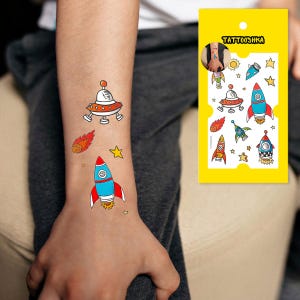 May include: A sheet of temporary tattoos featuring a variety of colorful space-themed designs, including rockets, planets, and stars. The sheet is labeled "Tattooshka".