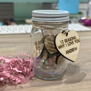 May include: A glass jar filled with small wooden hearts, each with a handwritten message. A larger wooden heart with the text "12 REASONS WHY I LOVE YOU ANDREW" is attached to the jar.