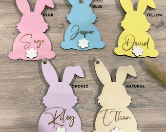 May include: Six wooden bunny ornaments in various colors, including pink, blue, yellow, orchid, and natural. Each bunny has a name written on it, including Sage, Jasper, David, Riley, and Ethan. The bunnies are all facing forward with their ears up.