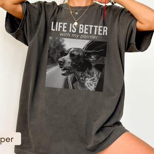 Op de afbeelding: Een antracietgrijs t-shirt met korte mouwen en een zwart-witte afbeelding van een hond met een zonnebril die uit een autoraam kijkt. Boven de afbeelding staat de tekst "LIFE IS BETTER with my pointer" in witte hoofd- en kleine letters.