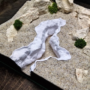 May include: A miniature terrarium scene with a sandy base, white rock formations, and small green plants. A white, flowing element resembles a river or path. The scene is enclosed in a glass container, creating a small-scale landscape.