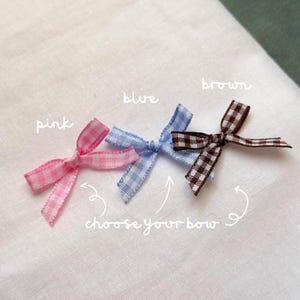 May include: Three small gingham bows in pink, blue, and brown are displayed on a white surface. The bows are tied with thin ribbon and have a classic bow shape. The text "choose your bow" is written below the bows.