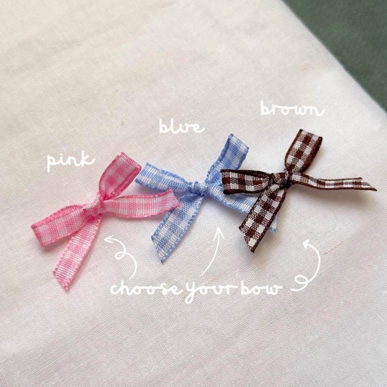 May include: Three small gingham bows in pink, blue, and brown are displayed on a white surface. The bows are tied with thin ribbon and have a classic bow shape. The text "choose your bow" is written below the bows.