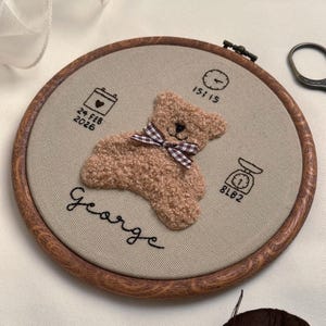 May include: Embroidered hoop art featuring a plush teddy bear with a checkered bow, the name "George", and birth details: date, time, and weight (8 lbs 2 oz). The design is set in a wooden hoop.