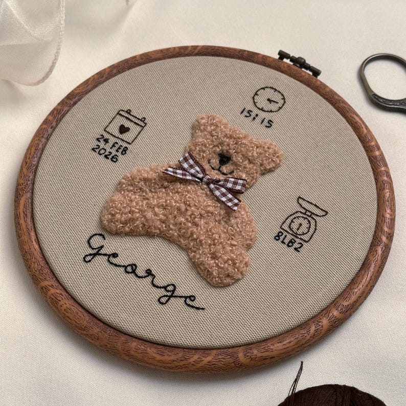 May include: Embroidered hoop art featuring a plush teddy bear with a checkered bow, the name "George", and birth details: date, time, and weight (8 lbs 2 oz). The design is set in a wooden hoop.