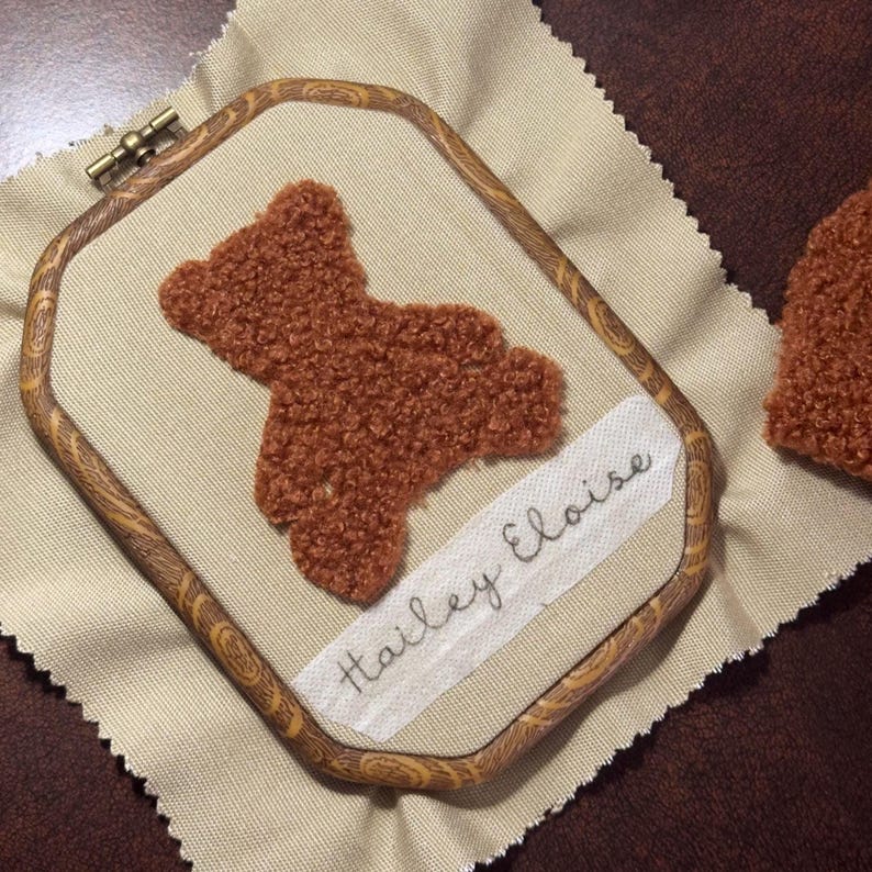 May include: A brown teddy bear embroidery in a wooden hoop. The bear is made of textured fabric and is stitched onto a beige fabric. The name "Hailey Eloise" is embroidered on a white strip of fabric.