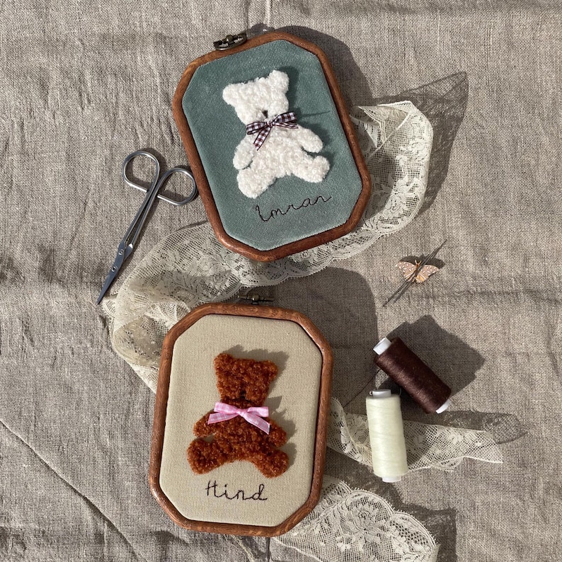 May include: Two framed embroidered teddy bears, one white with a checkered bow and the name "Imran", the other brown with a pink bow and the name "Hind". The frames are wooden with a teal and beige background. Scissors, thread, and lace are also visible.