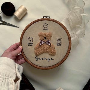 May include: A handmade, embroidered nursery hoop art featuring a teddy bear with a checkered bow tie. The design includes the name "George", birth date, time, and weight (8 lbs 2 oz). The hoop is made of wood and the fabric is beige.