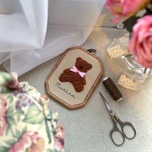 May include: A framed, embroidered teddy bear with a pink bow, the name "Marina" is stitched below. The bear is brown, set against a beige background, and framed in a wooden oval. Crafting supplies are nearby.