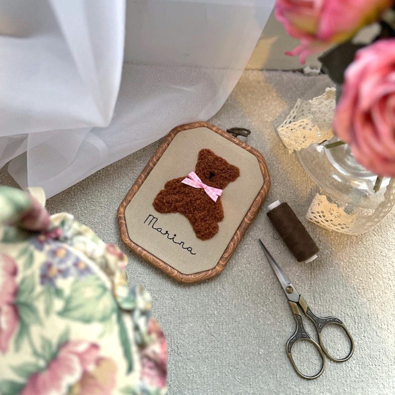 May include: A framed, embroidered teddy bear with a pink bow, the name "Marina" is stitched below. The bear is brown, set against a beige background, and framed in a wooden oval. Crafting supplies are nearby.