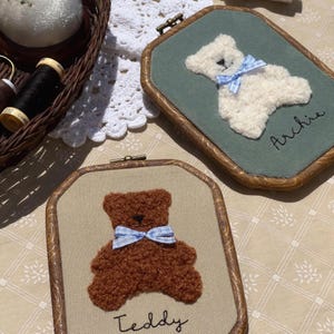 May include: Two framed embroidery pieces featuring teddy bears. One bear is white with a blue bow on a green background, the other is brown with a blue bow on a tan background. The names "Archie" and "Teddy" are embroidered below the bears.