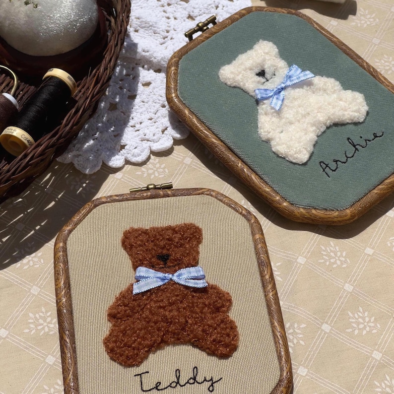 May include: Two framed embroidery pieces featuring teddy bears. One bear is white with a blue bow on a green background, the other is brown with a blue bow on a tan background. The names "Archie" and "Teddy" are embroidered below the bears.