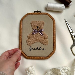 May include: A framed, embroidered teddy bear with a brown wooden frame. The bear is light brown with a checkered bow tie. The name "freddie" is embroidered below the bear in cursive. The frame is approximately 6 inches tall and 4 inches wide.