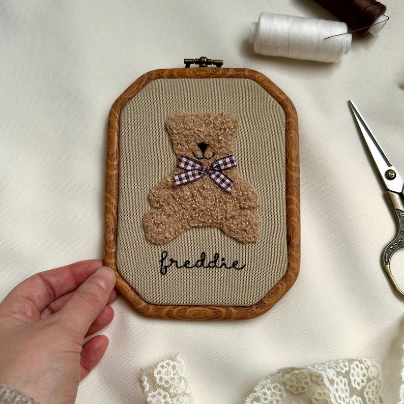 May include: A framed, embroidered teddy bear with a brown wooden frame. The bear is light brown with a checkered bow tie. The name "freddie" is embroidered below the bear in cursive. The frame is approximately 6 inches tall and 4 inches wide.
