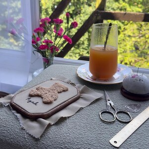 May include: An embroidery hoop with a beige teddy bear design and the name "Ayah" stitched in black thread. A glass of orange juice sits on a floral saucer. Other items include a pincushion, scissors, and a ruler.