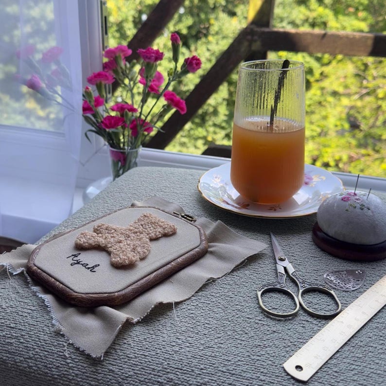 May include: An embroidery hoop with a beige teddy bear design and the name "Ayah" stitched in black thread. A glass of orange juice sits on a floral saucer. Other items include a pincushion, scissors, and a ruler.