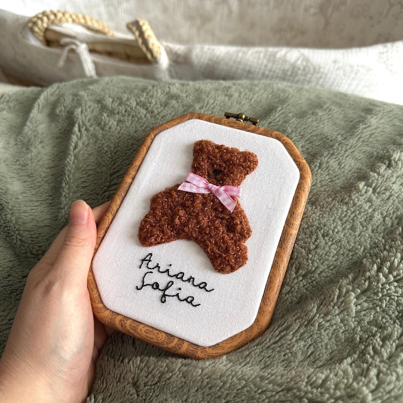 May include: A brown teddy bear embroidery on a white background, framed in a wooden hoop. The bear has a pink and white checkered bow. The name "Ariana Sofia" is embroidered below the bear in black script.