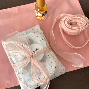 May include: A wrapped gift with a pink ribbon and a small bottle of perfume on a pink surface. The gift is wrapped in white paper with colorful confetti and a tag that says "thank you". A roll of pink ribbon is also visible.