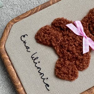 May include: A brown teddy bear embroidery on a beige fabric, framed in a light brown wooden hoop. The bear has a pink bow. The name "Ena Winnie" is embroidered in black cursive on the fabric.