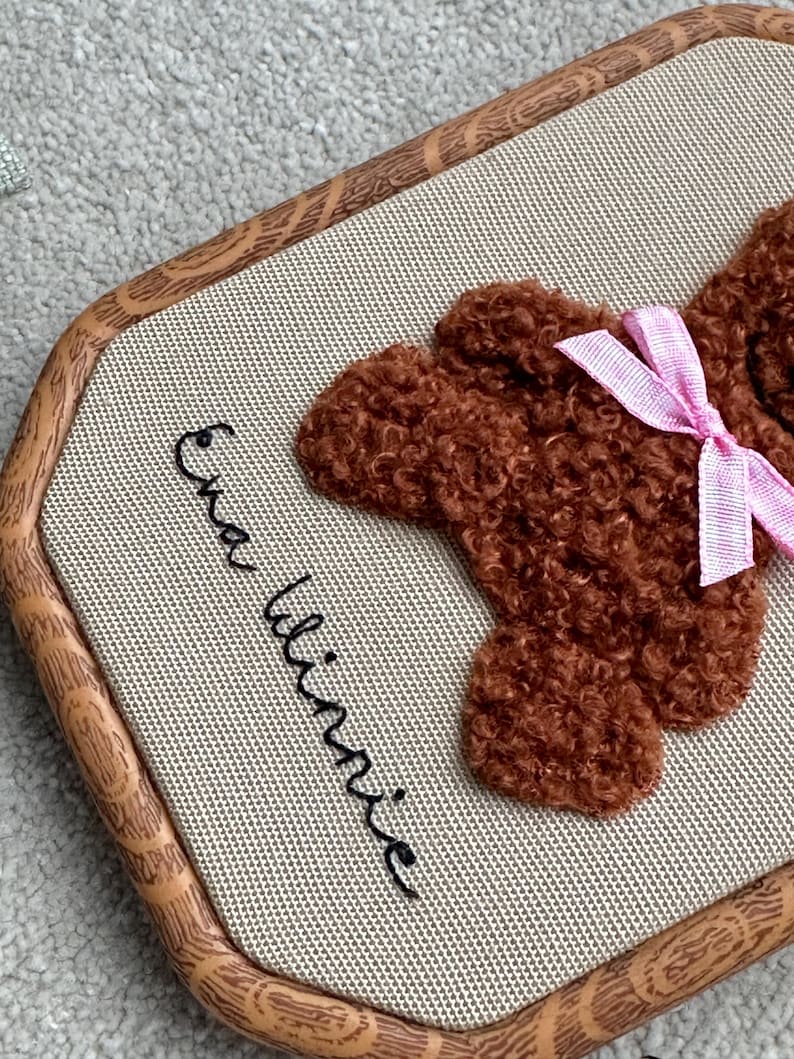 May include: A brown teddy bear embroidery on a beige fabric, framed in a light brown wooden hoop. The bear has a pink bow. The name "Ena Winnie" is embroidered in black cursive on the fabric.