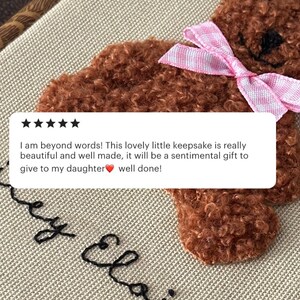 May include: A brown teddy bear with a pink and white bow, embroidered on a beige fabric. The image includes a customer review with five stars and the text: "I am beyond words! This lovely little keepsake is really beautiful and well made..."