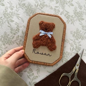May include: A framed, embroidered artwork featuring a brown teddy bear with a blue gingham bow. The name "Rowan" is stitched below the bear. The frame has a wood-grain appearance. A pair of scissors and a piece of brown felt are in the background.