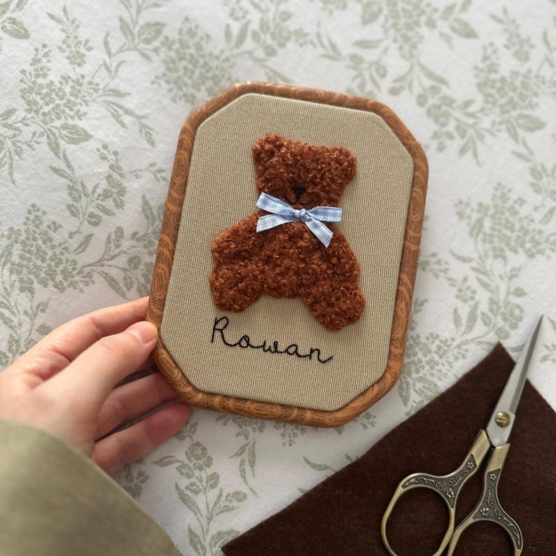 May include: A framed, embroidered artwork featuring a brown teddy bear with a blue gingham bow. The name "Rowan" is stitched below the bear. The frame has a wood-grain appearance. A pair of scissors and a piece of brown felt are in the background.