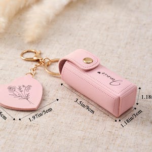 May include: A light pink lipstick case with a gold snap closure and the name Jennie printed on the side. It includes a heart-shaped keychain charm with a floral line drawing. The case measures 3.5 inches by 1.18 inches, and the charm is 1.97 inches.