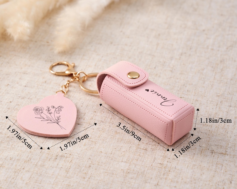May include: A light pink lipstick case with a gold snap closure and the name Jennie printed on the side. It includes a heart-shaped keychain charm with a floral line drawing. The case measures 3.5 inches by 1.18 inches, and the charm is 1.97 inches.