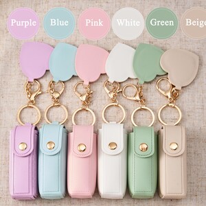 May include: Six rectangular lipstick cases with snap closures, each featuring a heart-shaped charm attached to a gold-toned metal keyring. The cases come in purple, blue, pink, white, green, and beige, with corresponding color labels above each.