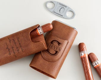 May include: A brown leather cigar case and two cigars with white and orange bands. The case is embossed with the name "STEVEN" and a large initial. A silver cigar cutter with the name "JASMINE" and the word "Groomsman" is also present.