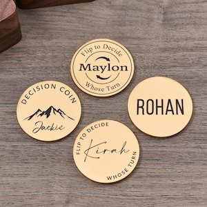 May include: Four gold-coloured decision coins with black text. One coin reads "Maylon" with "Flip to Decide" and "Whose Turn". Another says "Jackie" with a mountain graphic and "Decision Coin". The third coin says "Rohan". The fourth coin says "Kirah" with "Flip to Decide" and "Whose Turn".