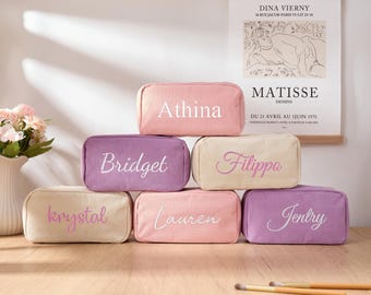 May include: Six personalized makeup bags in pink and purple corduroy fabric. The bags are stacked in a pyramid shape and feature the names Athina, Bridget, Filippo, Krystal, Lauren, and Jentry embroidered in white.