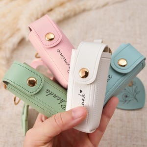 May include: A collection of four small, rectangular lipstick cases with gold-tone snap closures. The cases are available in pastel pink, sage green, white, and light blue, each featuring personalized names and floral line art printed in black ink.