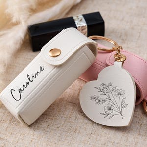 May include: A white lipstick case with a gold snap closure and the name Caroline printed in black script. Attached to the case is a heart-shaped white leather keychain featuring a black line drawing of a floral bouquet. A pink case is nearby.